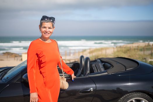 Stylish woman in orange dress enjoys a sunny day by the beach with a sleek black convertible in Portugal.
