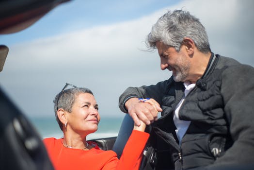 Smiling couple holding hands and looking at each other by the ocean, showcasing love and connection.