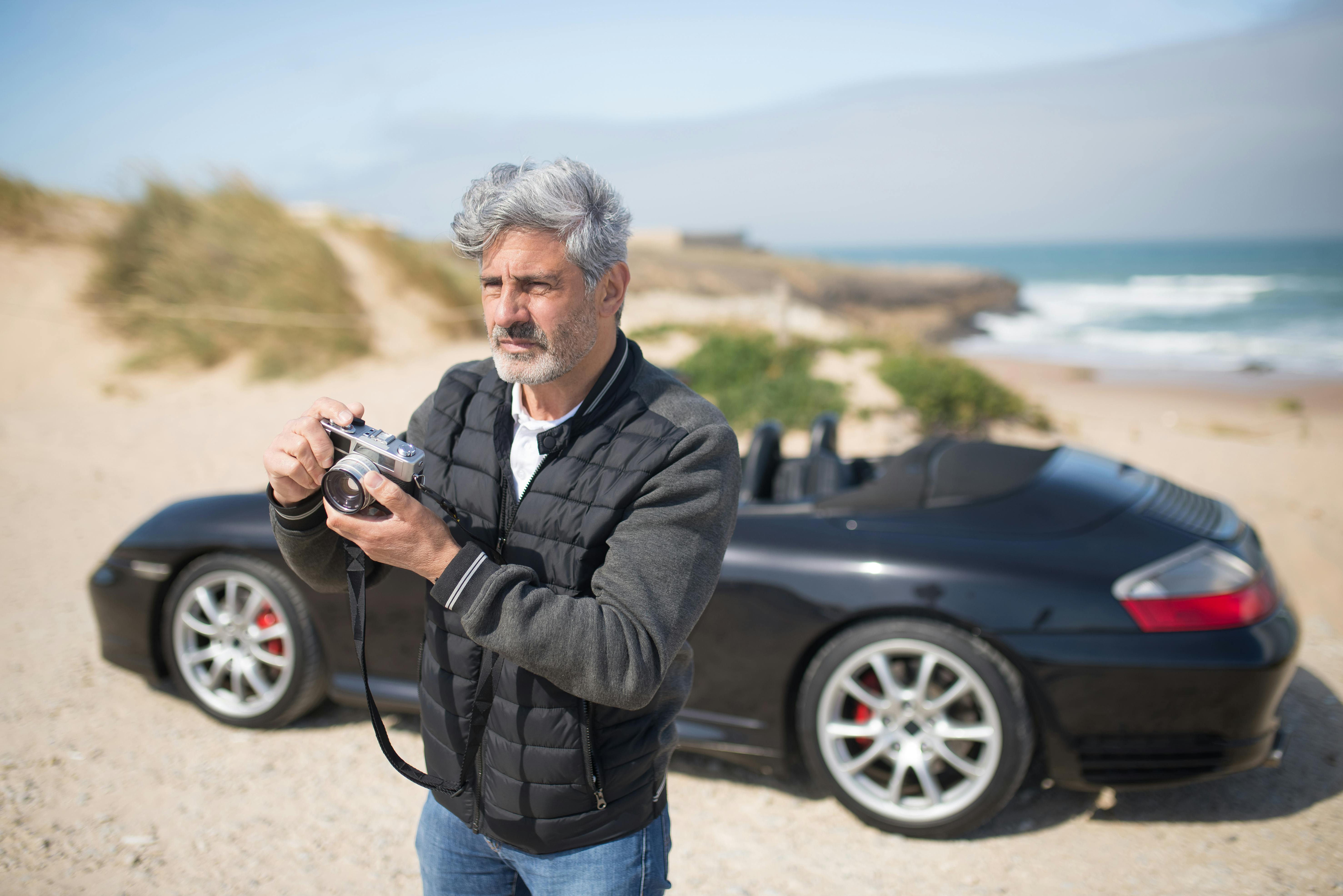 Senior man holding a camera by a convertible car on a Portugal beach, capturing the scenic view.