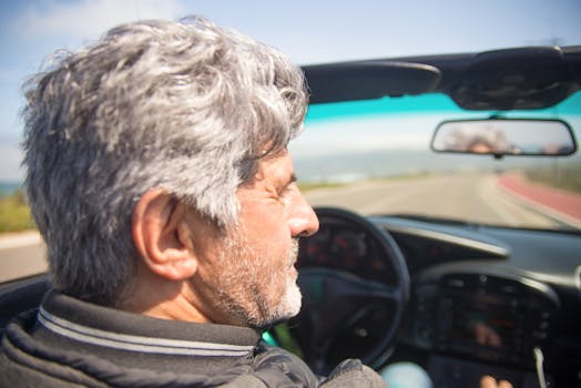 An elderly man with gray hair enjoys a sunny drive in a convertible along a scenic road.