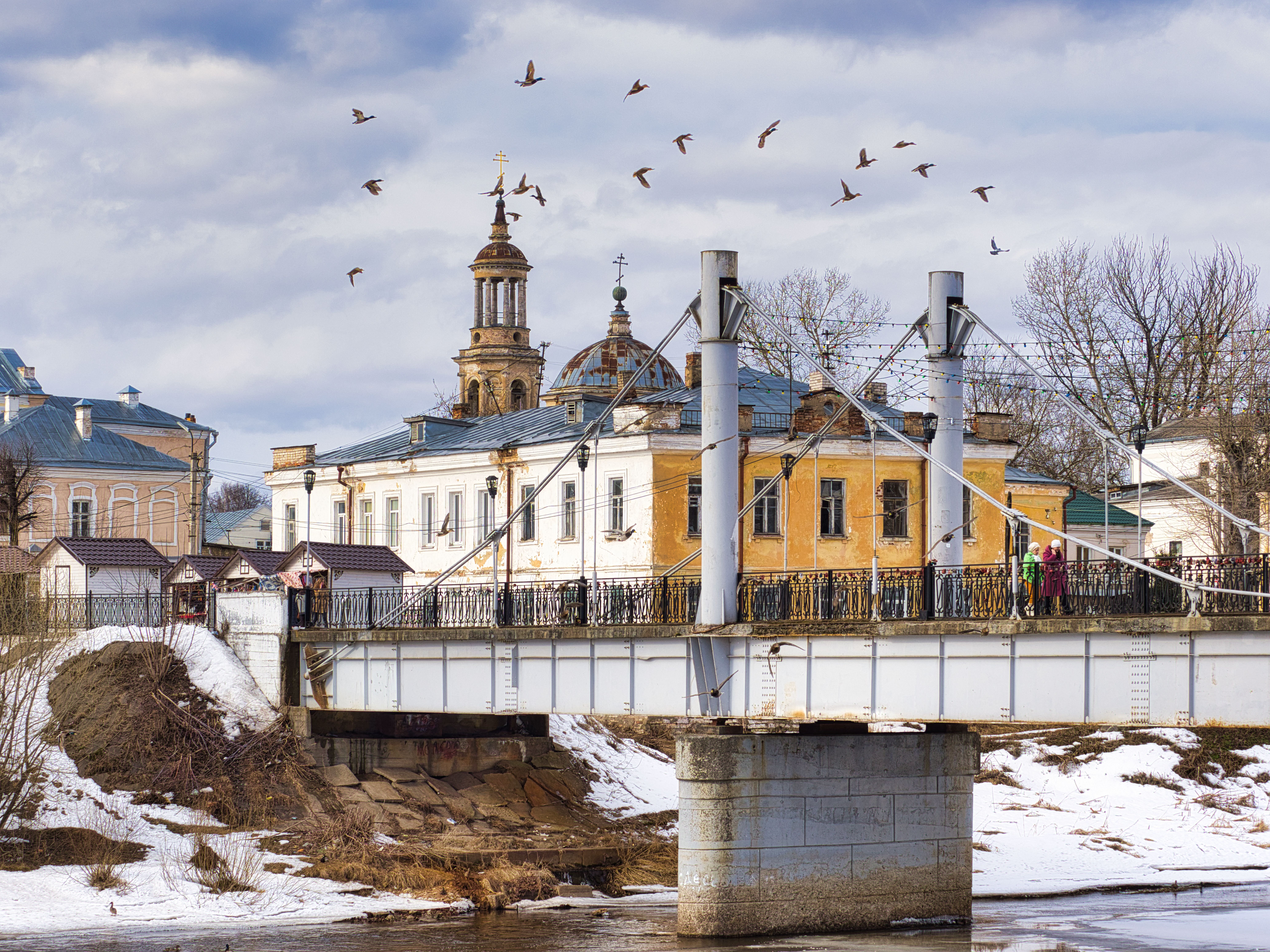 A scenic view of a historic bridge and church in winter, Торжок, Russia, with birds flying overhead.