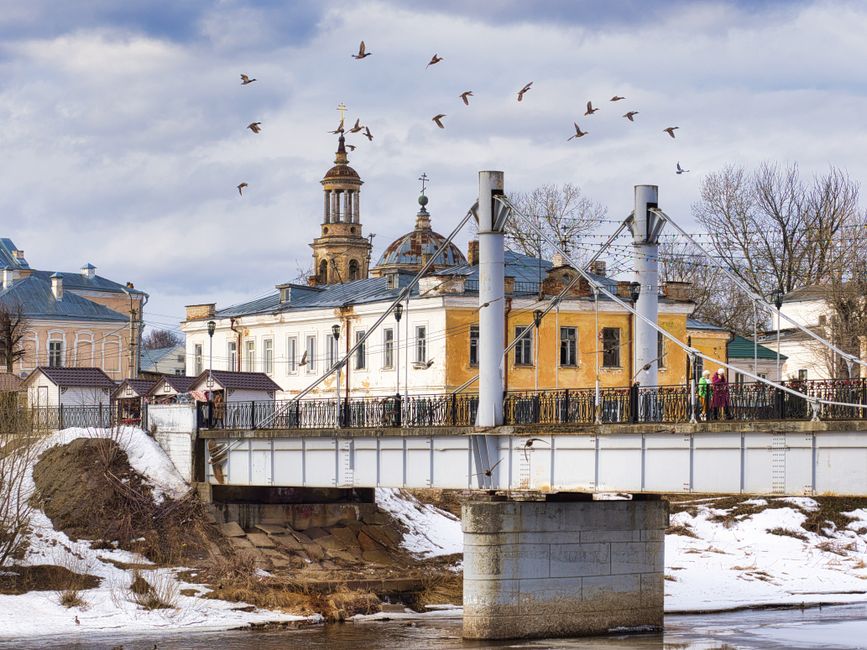 A scenic view of a historic bridge and church in winter, Торжок, Russia, with birds flying overhead.