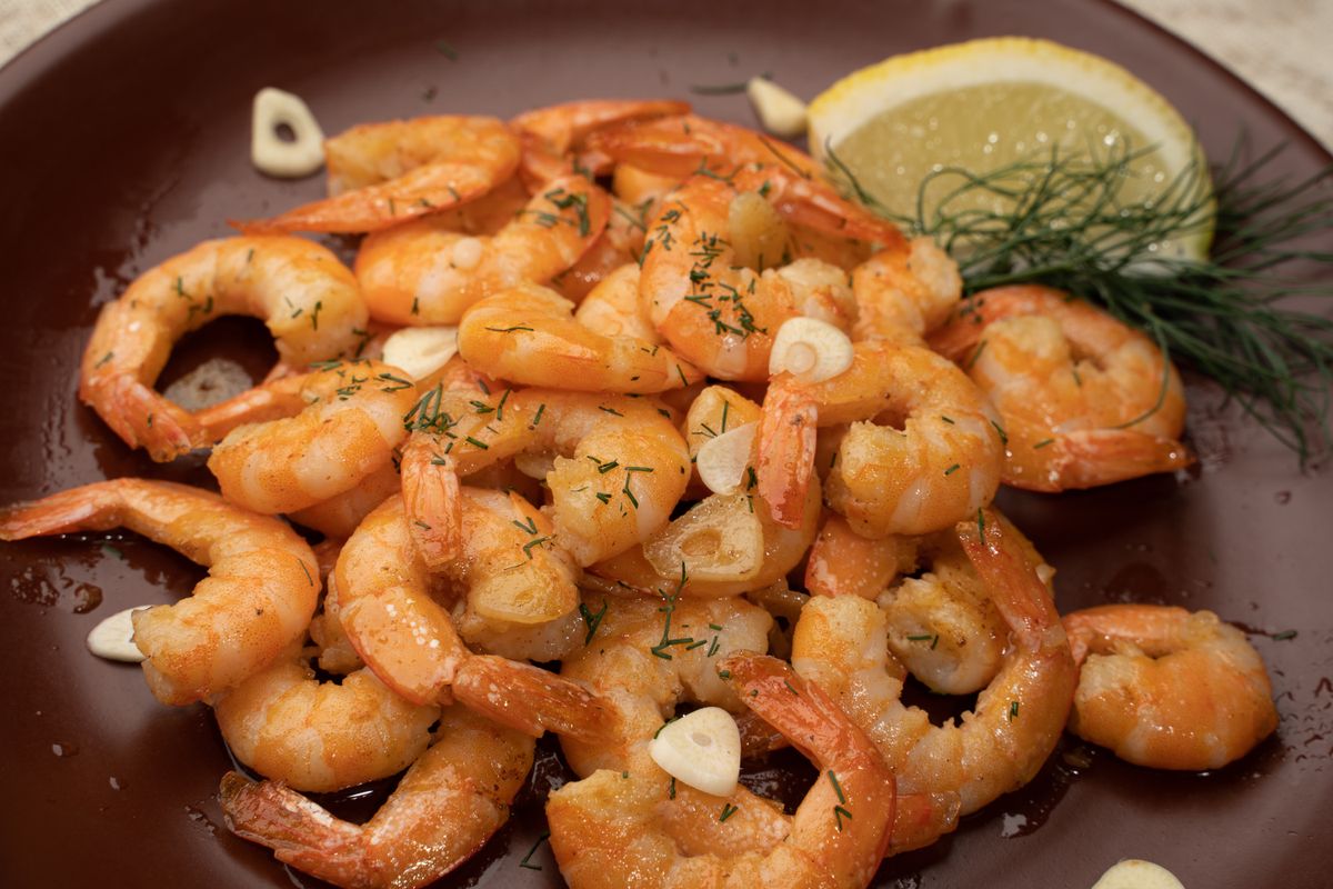 Sauteed pink shrimp with sliced garlic and herbs on a brown plate with lemon and dill