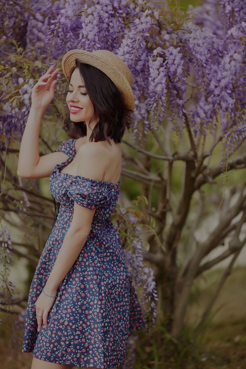 Free A young woman poses smiling in a blue floral dress and brown hat among vibrant purple wisteria blooms. Stock Photo
