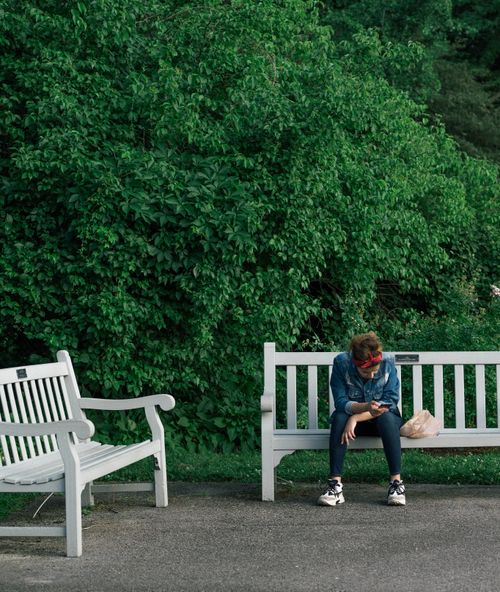 Free A young woman sitting on a white bench in a park, engrossed in her phone. Stock Photo