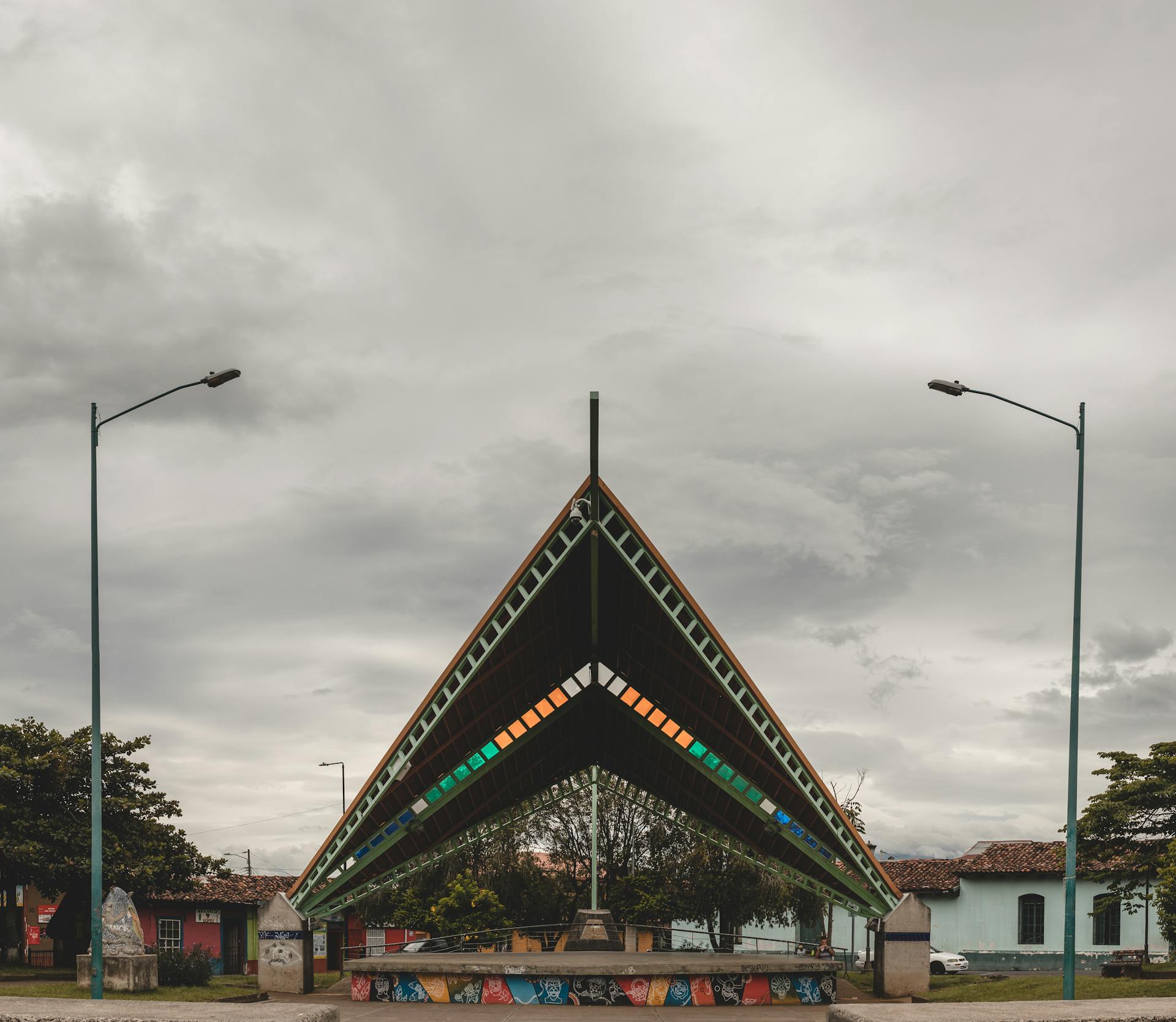 A modern pavilion with colorful roof panels under a cloudy sky surrounded by urban scenery.