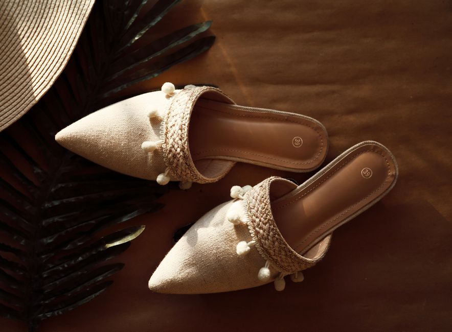 Stylish beige sandals with decorative pom-poms on a summer backdrop, featuring woven straw elements.