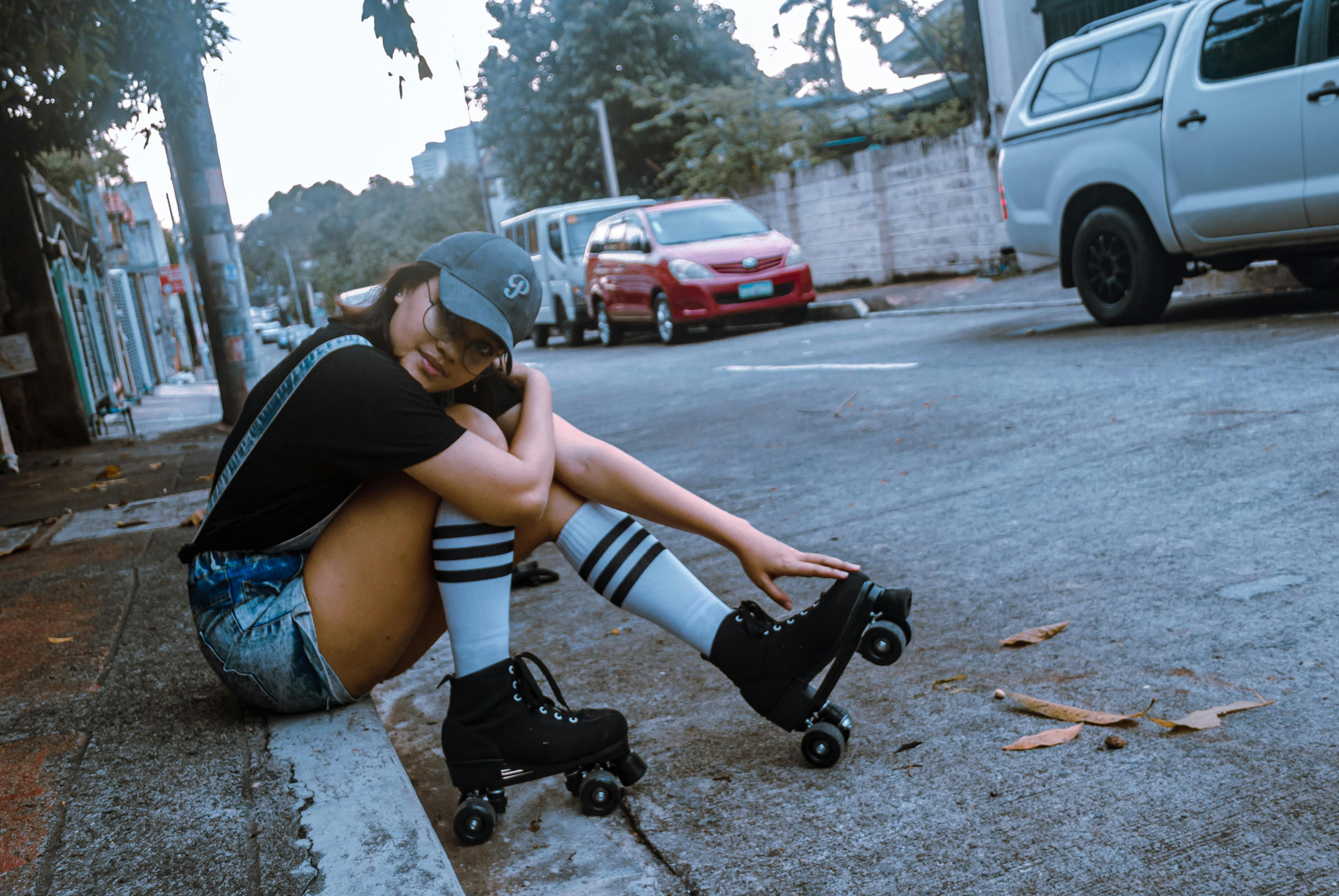 A young woman in roller skates sits on a curb on a lively urban street with cars parked nearby.