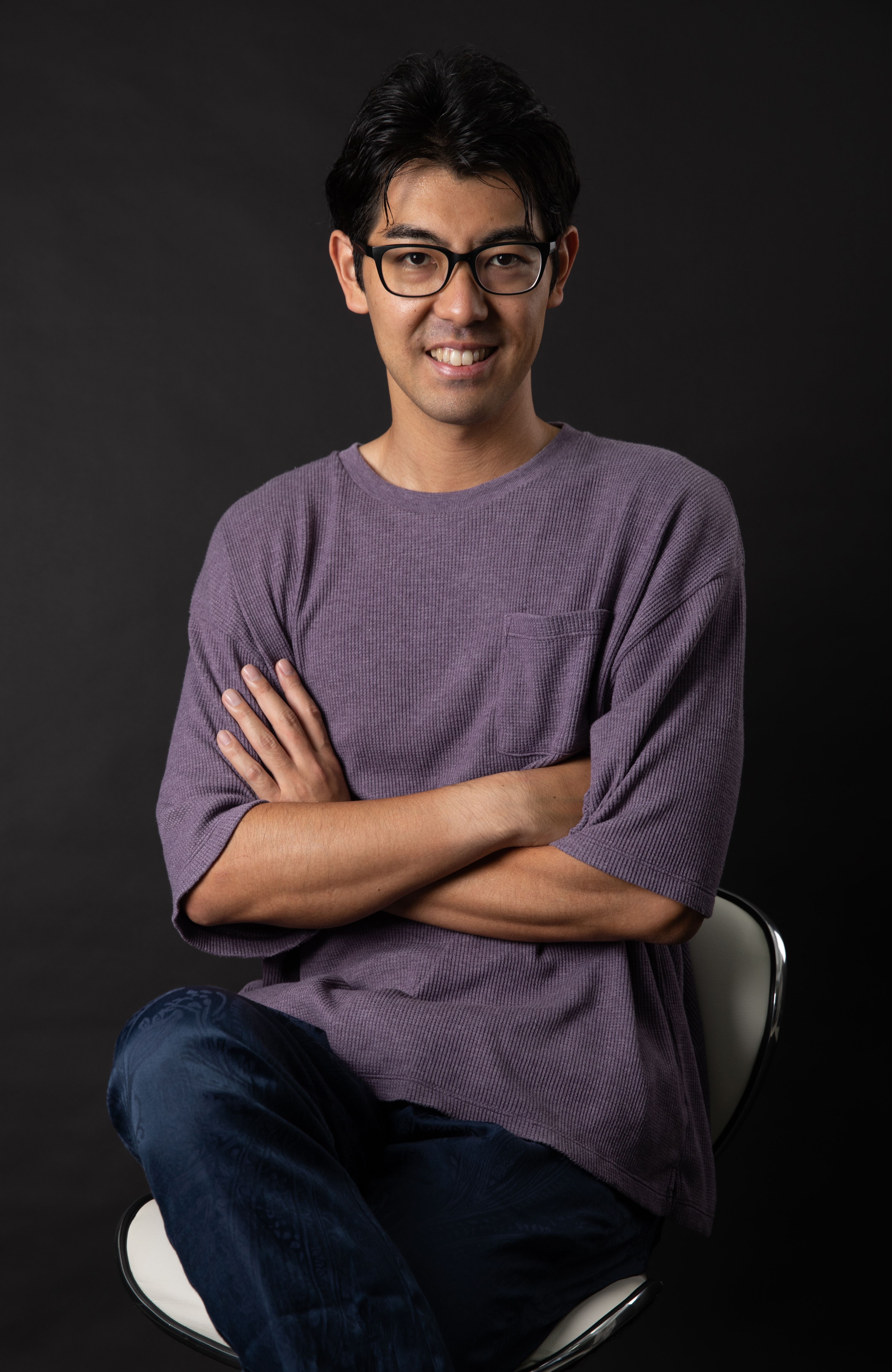 Casual portrait of a smiling man sitting with crossed arms against a black background.