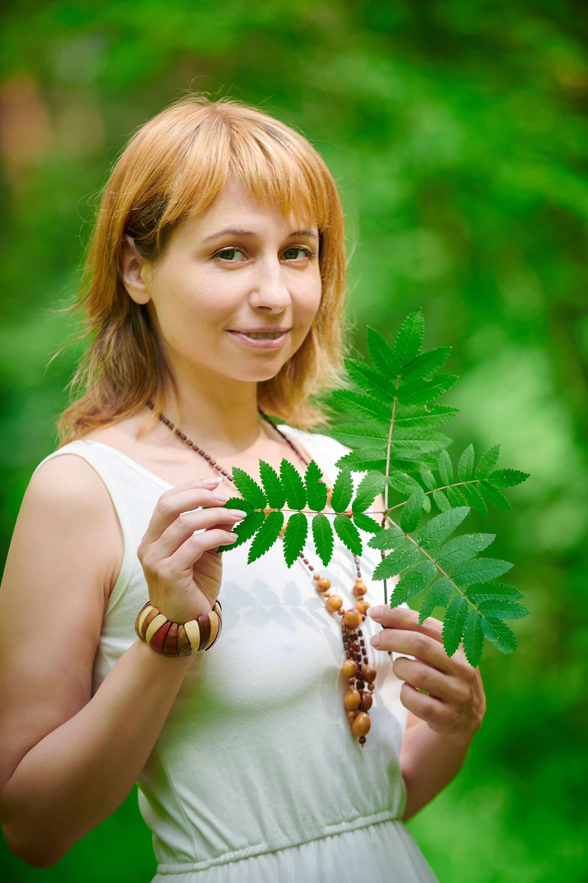 A woman holds vibrant green leaves while smiling in a lush outdoor setting.