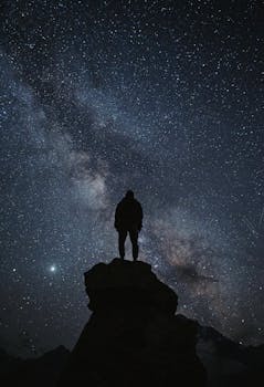 Person silhouette standing on rock under a vast starry sky with Milky Way, showcasing serene natural beauty.
