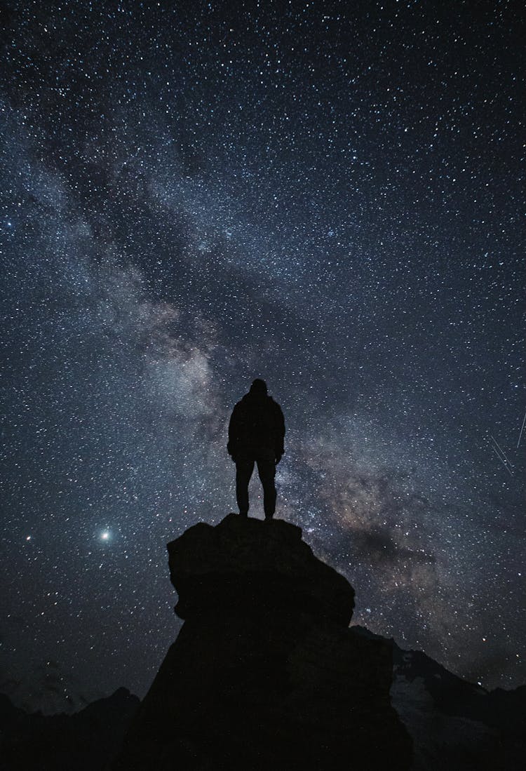 A Silhouette Of Person Standing On A Rock Under The Starry Sky