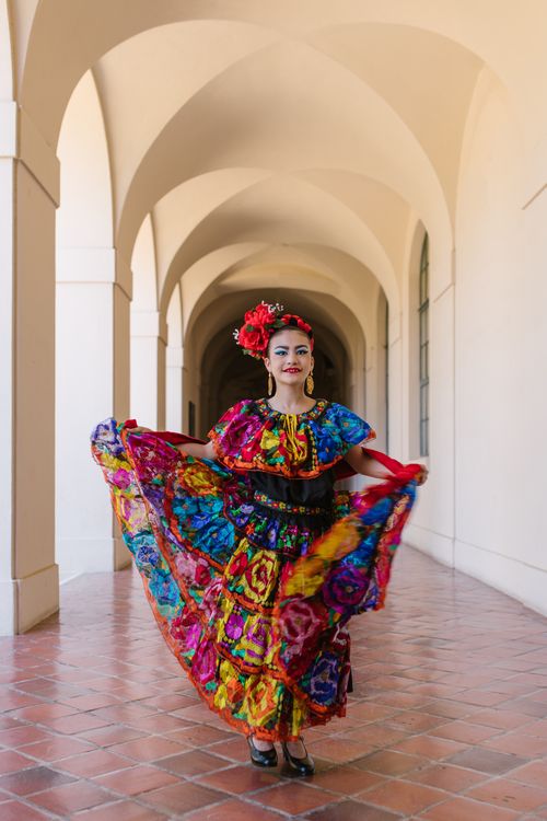Free Vibrant Mexican dancer in traditional dress with a colorful headdress in a scenic, arched hallway. Stock Photo