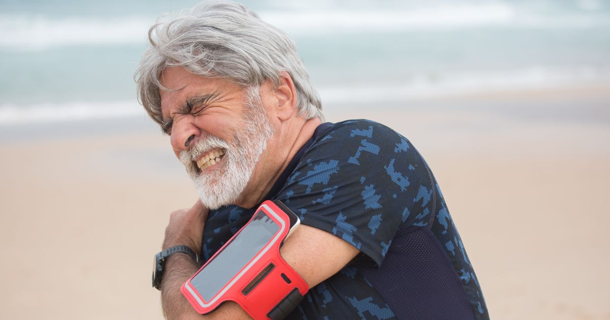 Senior man with gray hair and beard grimacing and holding his shoulder in pain