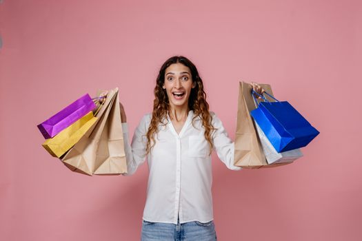 Smiling woman holding colorful shopping bags against a pink background, expressing excitement.