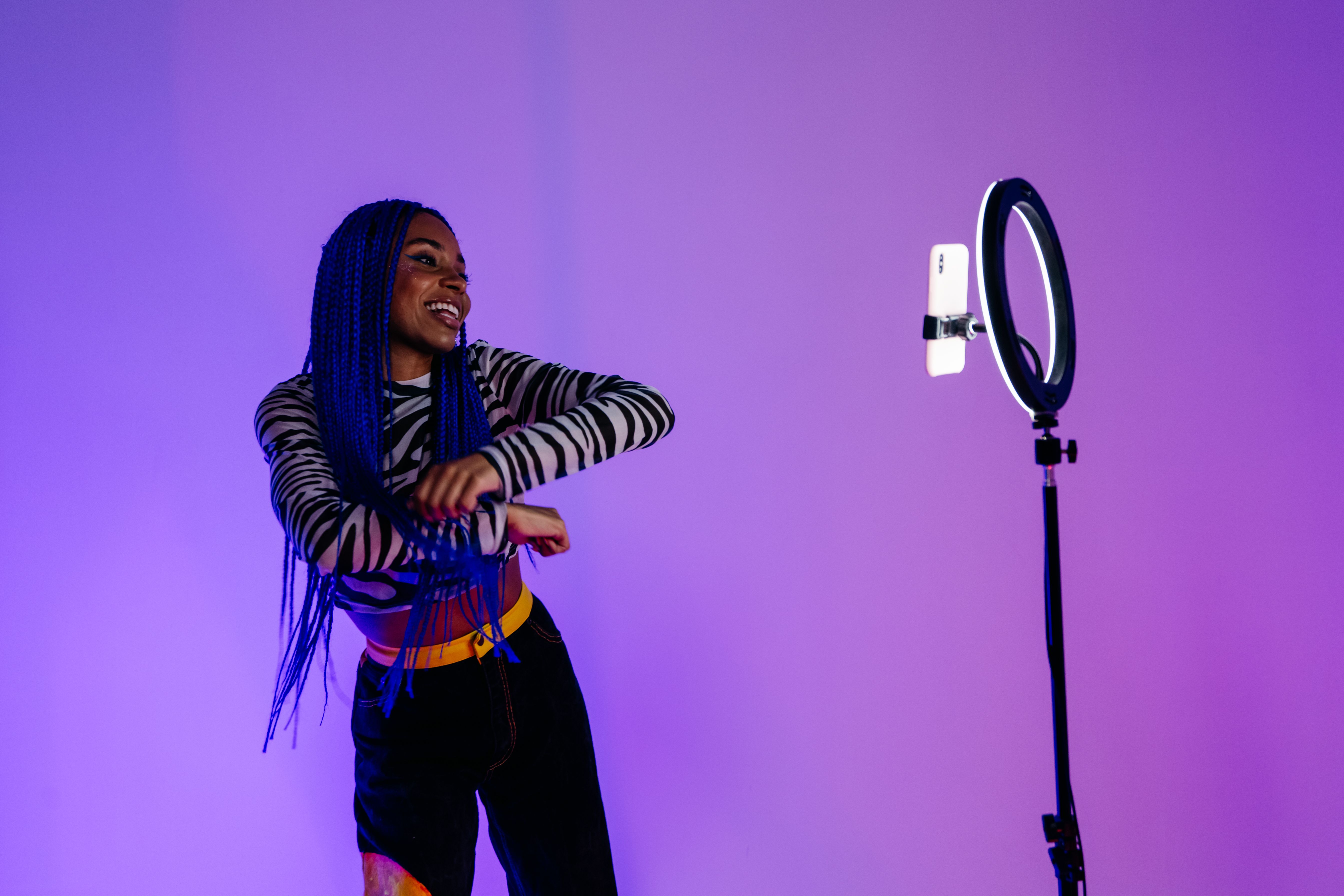 A young African American woman with braids dances joyfully in front of a ring light on a purple background.