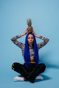 Fashionable woman with blue hair holding a pineapple on head against blue background. Indoor studio shoot.