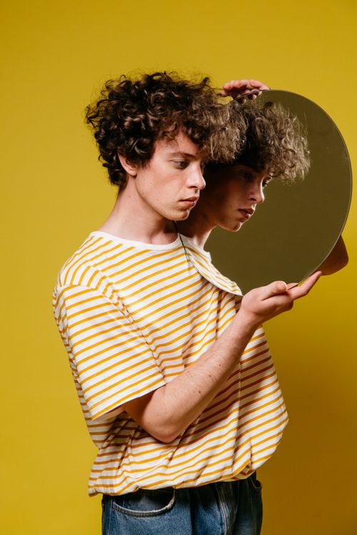 Free Stylish young man in striped shirt holding circular mirror against a vibrant yellow background. Stock Photo