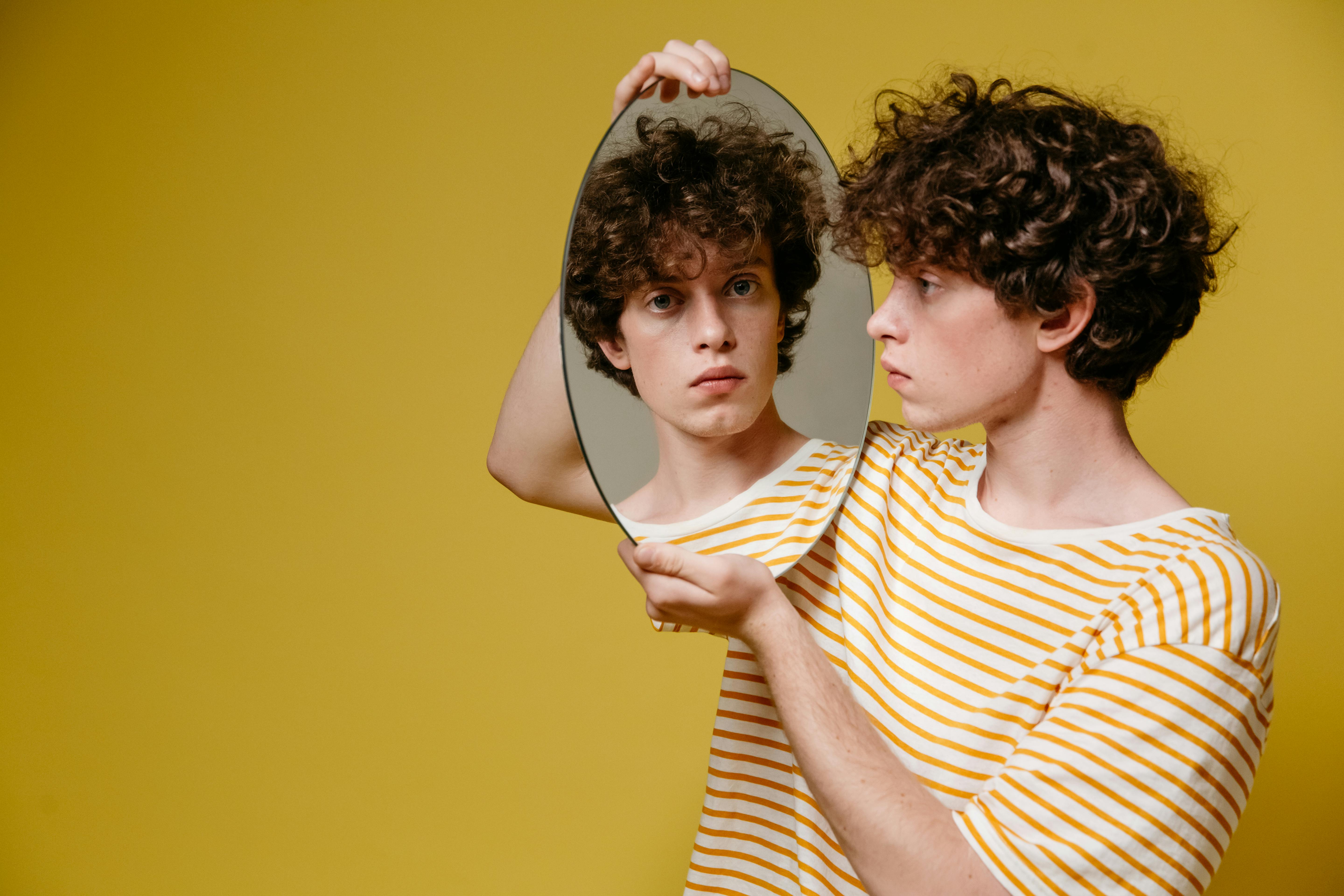 A young man with curly hair holds a mirror reflecting his serious expression against a yellow backdrop.