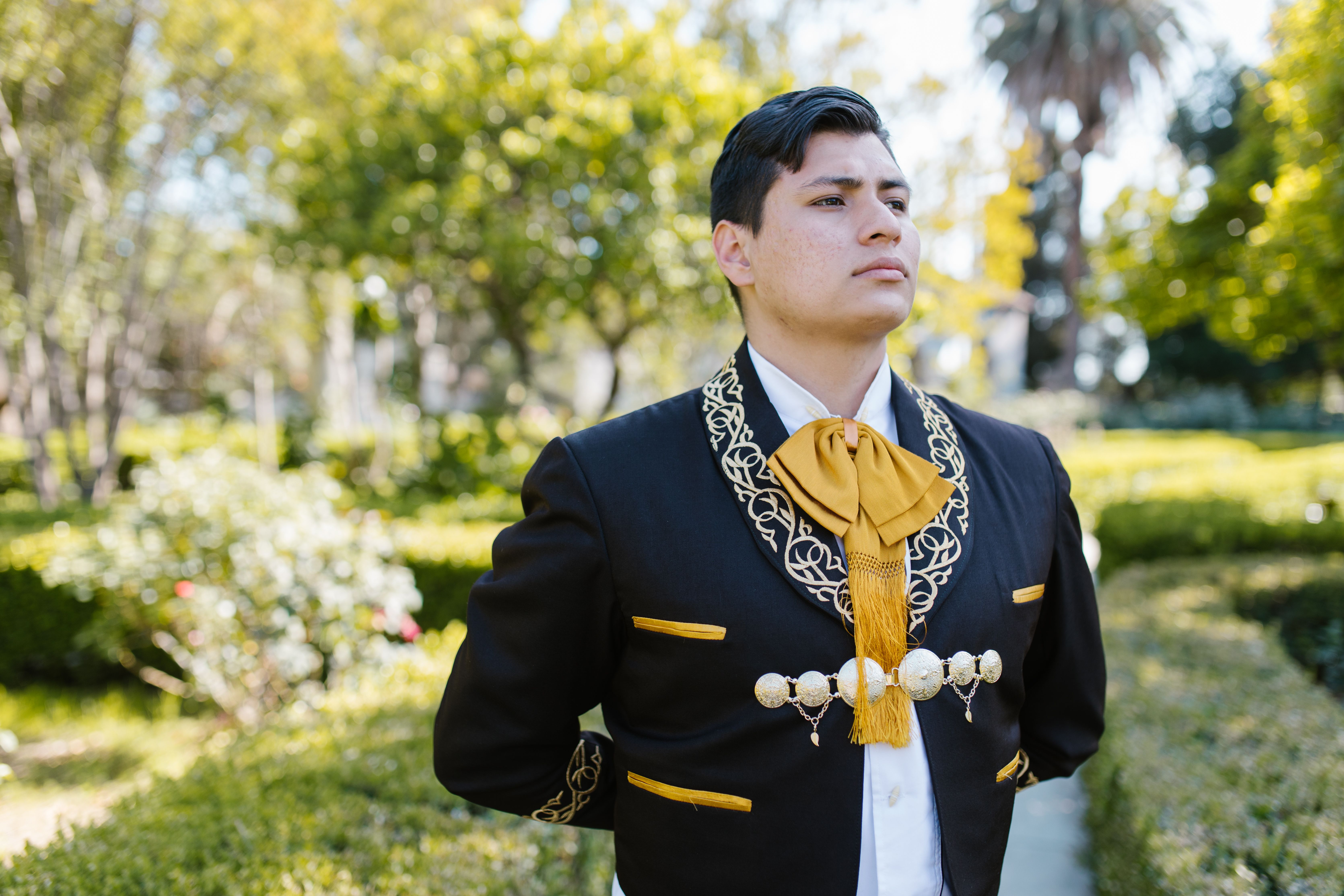 A man proudly wears a traditional charro suit in a lush, sunny garden.