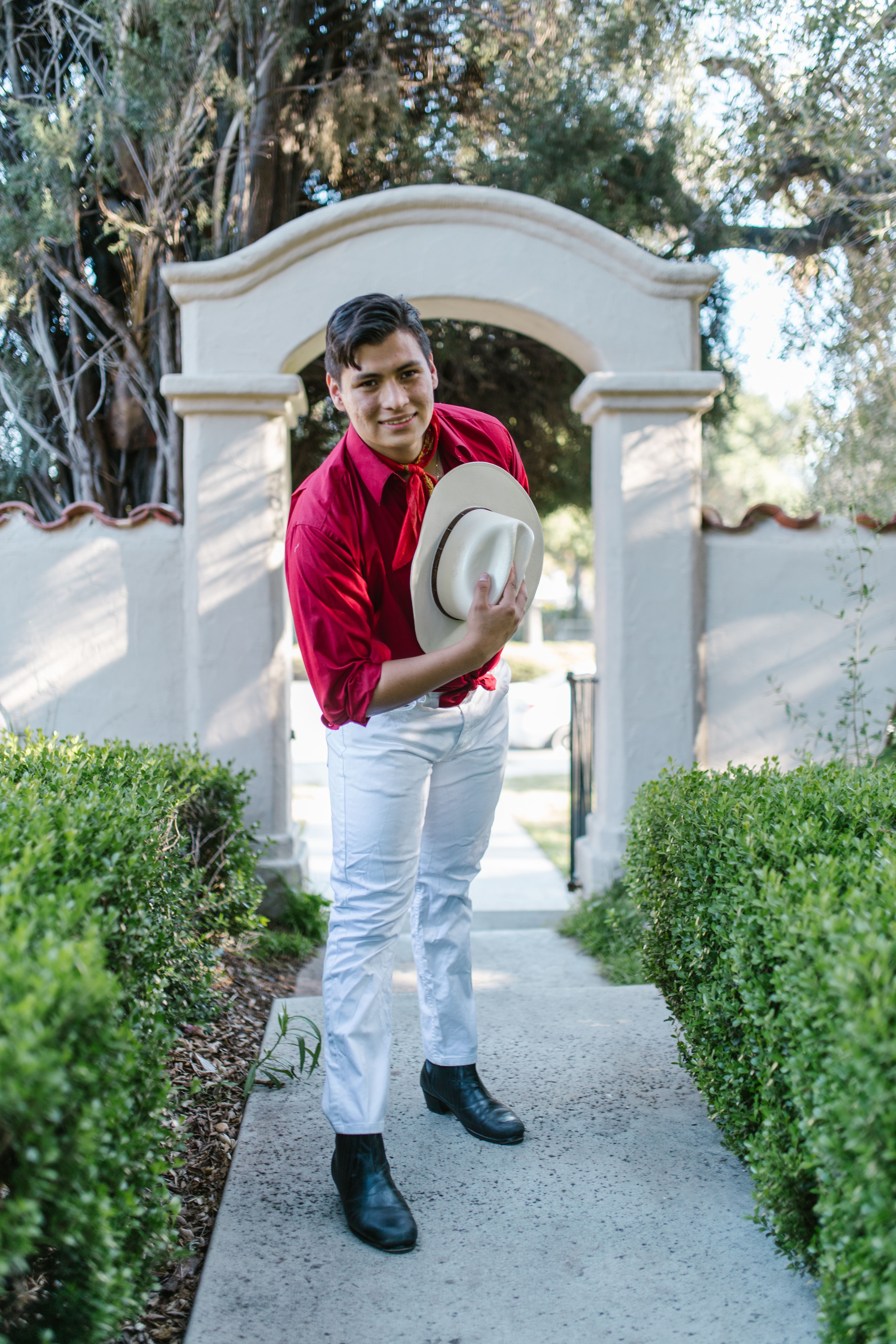 A cheerful man in traditional Mexican clothing, bowing with a hat in a garden setting.