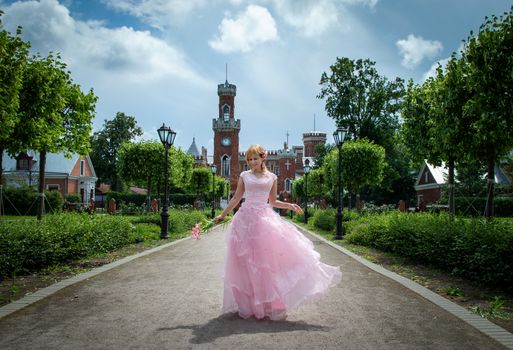A young woman in a pink dress walks gracefully along a garden path with a castle in the background.