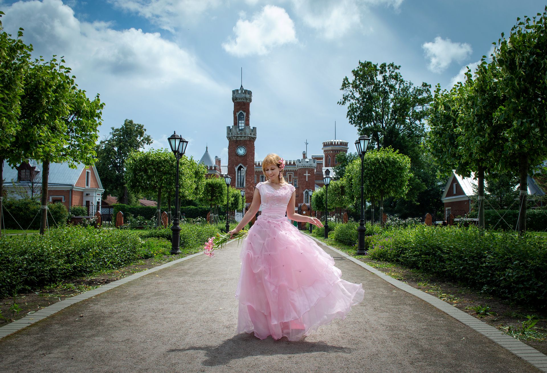 A young woman in a pink dress walks gracefully along a garden path with a castle in the background.