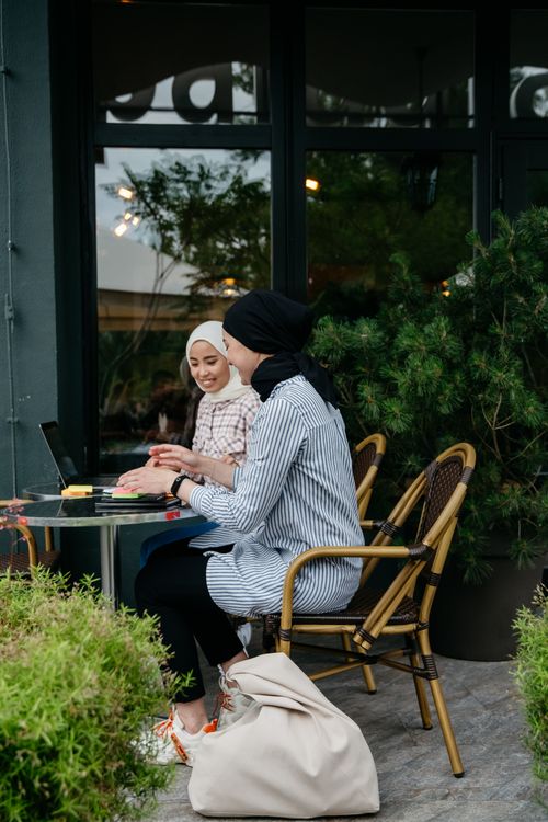 Free Two Muslim women in hijabs enjoying a discussion at an outdoor café, working on laptops surrounded by greenery. Stock Photo