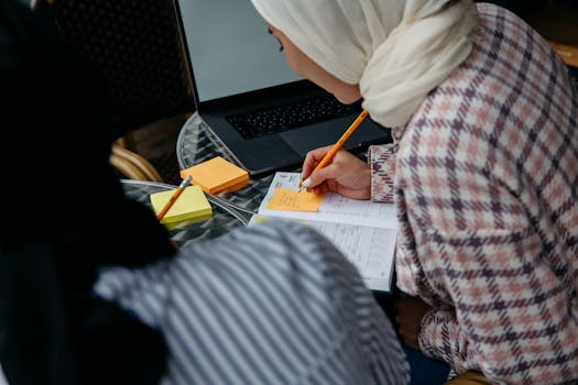 Muslim woman in hijab writing notes at café table with laptop and planner.