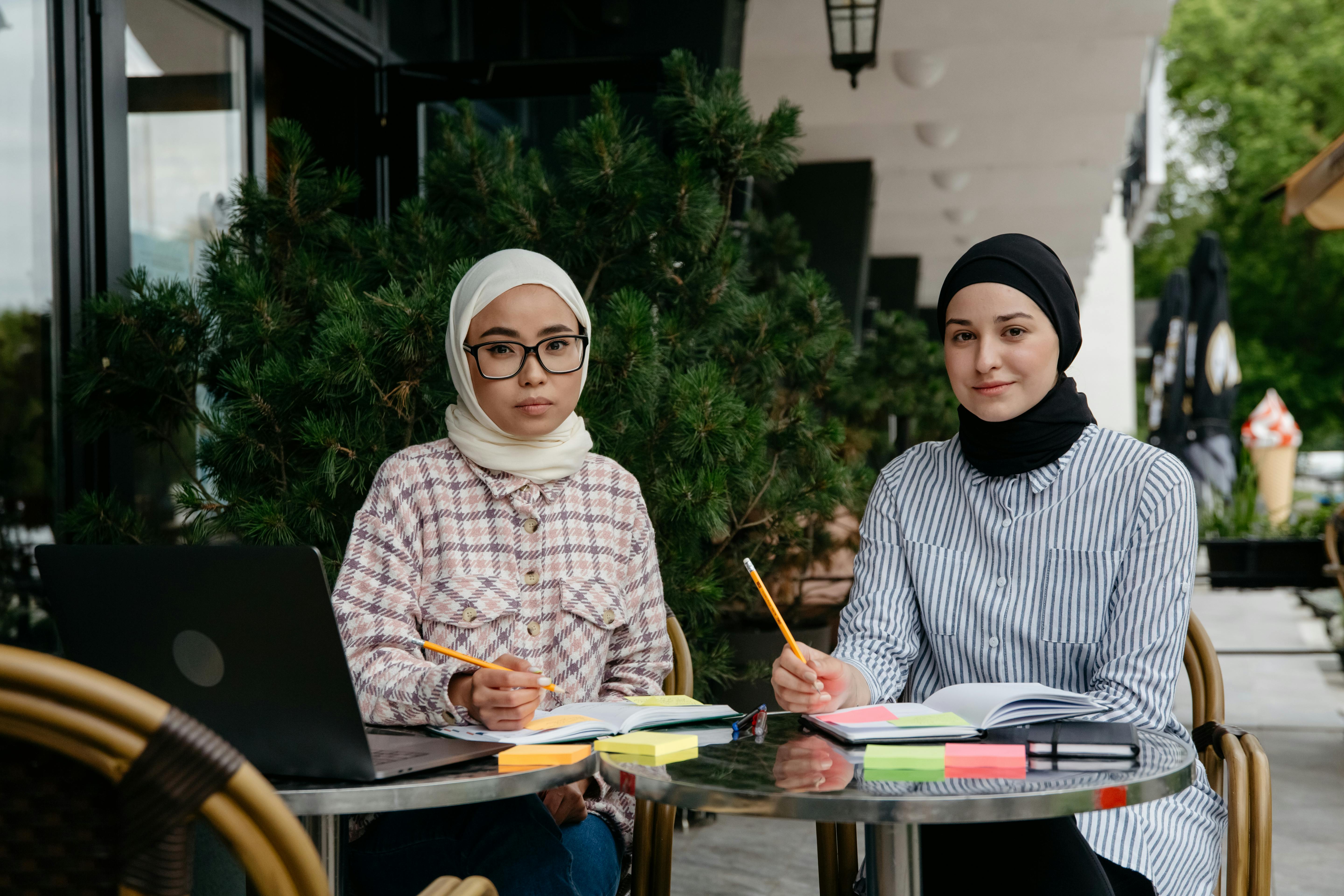 two women in white and black hijabs studying and writing on notebooks