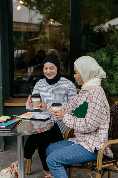 Free Two women in hijabs sharing coffee and studying at an outdoor café table in a relaxed atmosphere. Stock Photo