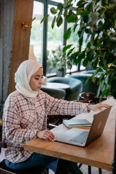 A young woman in hijab uses a laptop in a bright café, focusing on work or study.