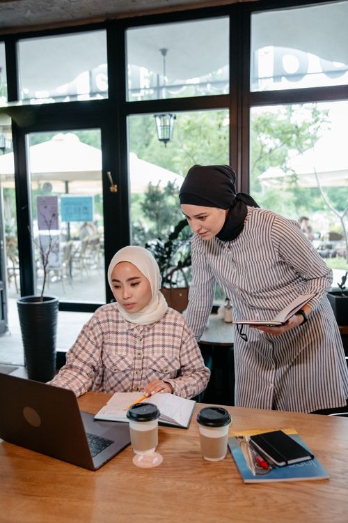 Free Two Muslim women wearing hijabs collaborate on a project using a laptop in a modern café. Stock Photo