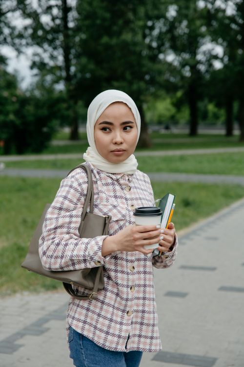 Free Young woman in a hijab holding coffee and a book, walking outdoors in a park setting. Stock Photo