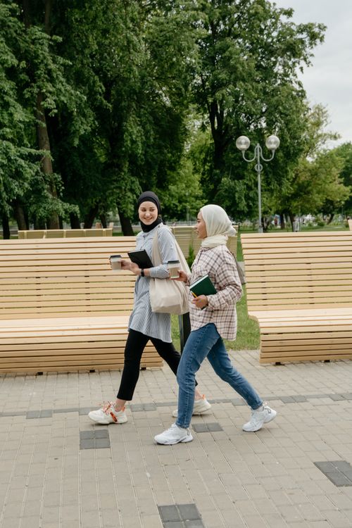 Free Two women in hijabs walking in a park, casually dressed and smiling, with benches and trees in the background. Stock Photo