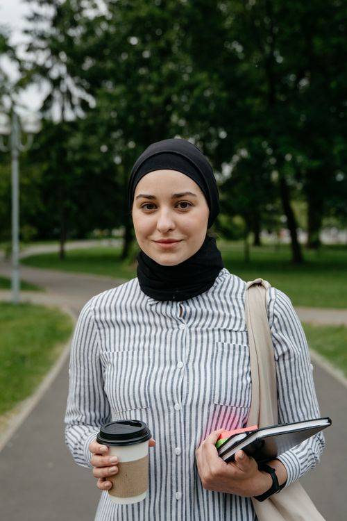 Free Smiling woman with hijab holding coffee and notebooks outdoors. Stock Photo