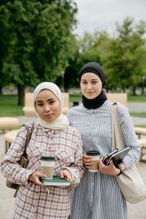 Free Two Muslim women in hijabs holding coffee cups and books in a park setting. Stock Photo