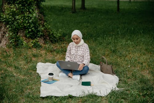 Woman in White Hijab Using Her Laptop while Siting on Picnic Blanket