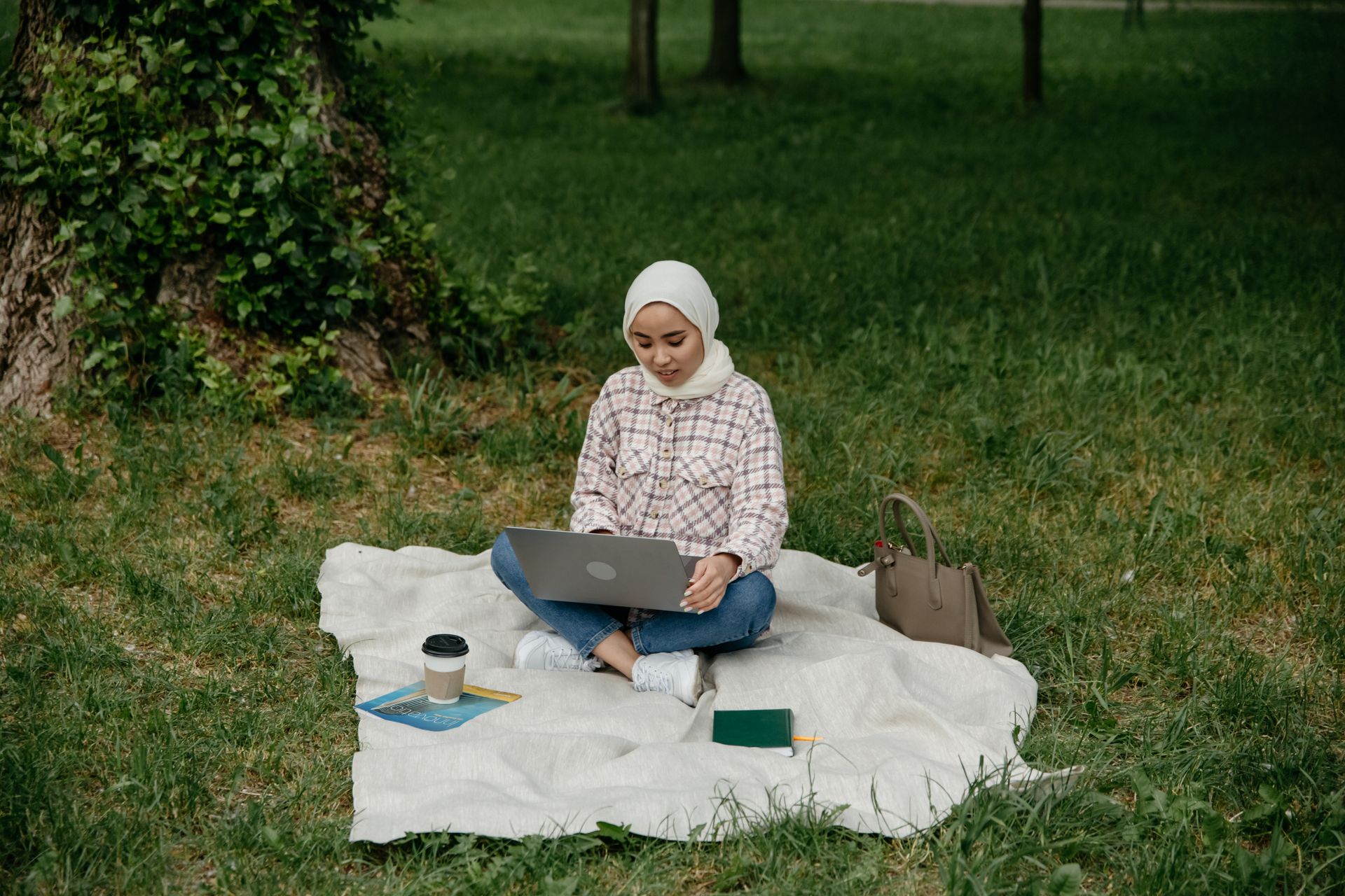 Young Asian woman in hijab using laptop on picnic blanket outdoors, enjoying nature and technology.