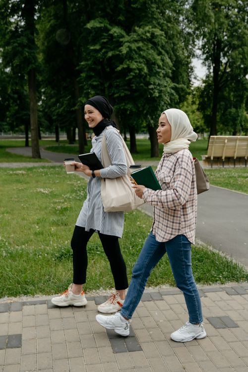 Free Two young Muslim women in hijabs walk through a green park, carrying books and coffee. Stock Photo