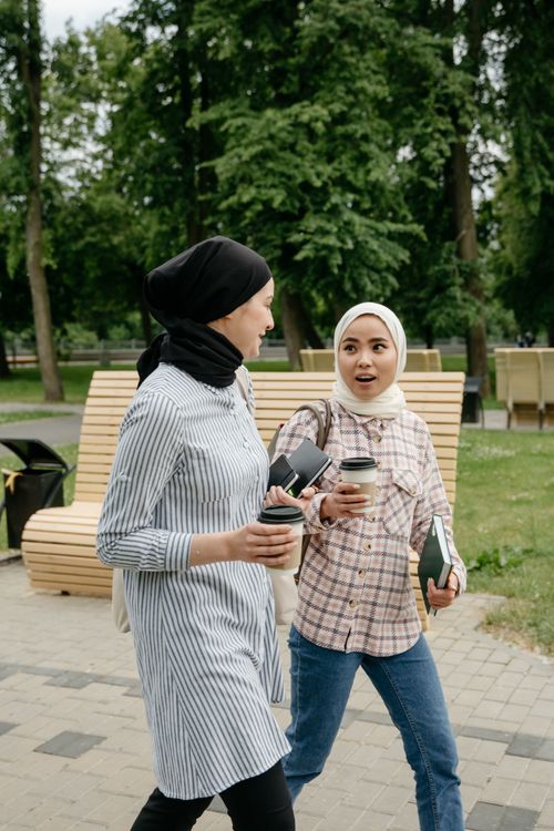 Free Two women in hijabs walking and chatting with coffee cups in a park setting. Stock Photo