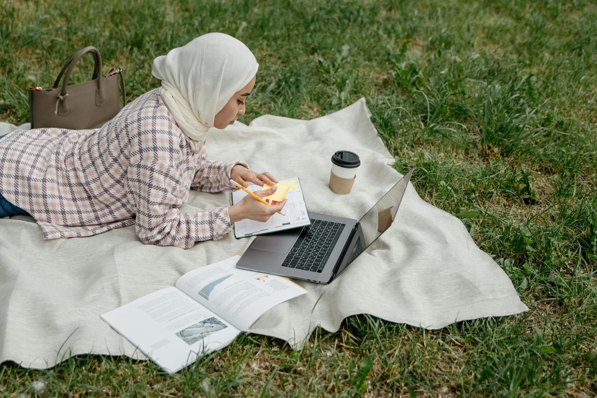 Muslim woman in hijab working on laptop outdoors on a picnic blanket.