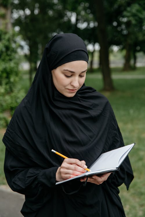 Woman in Black Hijab Writing on Notebook