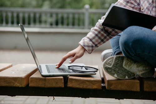 Person in Blue Denim Jeans and Plaid Shirt Sitting on Wooden Bench Using Laptop