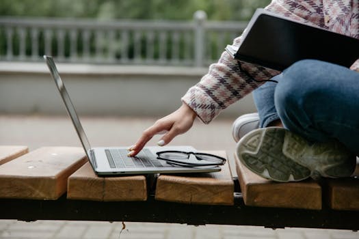 A person working on a laptop outdoors on a wooden bench, showcasing remote work in a natural setting.