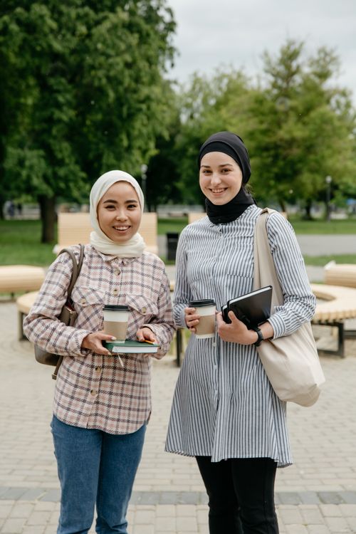 Free Two smiling young women wearing hijabs holding coffee cups and notebooks in a city park. Stock Photo