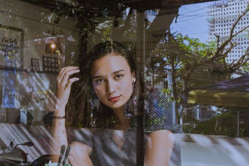Free A thoughtful young woman gazes through a café window, with reflections blending indoor and outdoor elements. Stock Photo