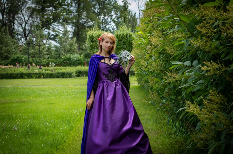 Woman in a purple gown posing in a lush green garden, exuding elegance and mystery.