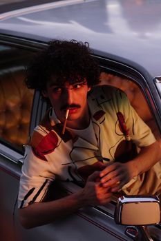 A fashionable man with curly hair smokes in a vintage car during sunset, creating a moody ambiance.