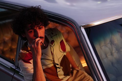 Free Man with curly hair and mustache smoking in a vintage car during twilight, creating a moody ambiance. Stock Photo